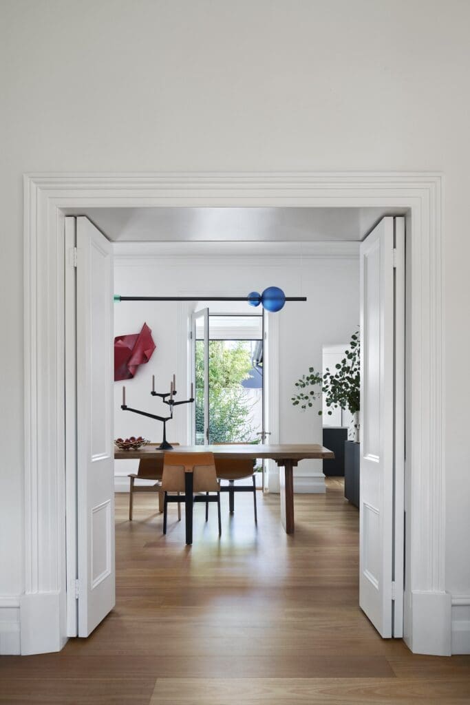 View through white double doors into a modern dining room with timber table, sculptural lighting and large window bringing natural light into the minimalist interior.