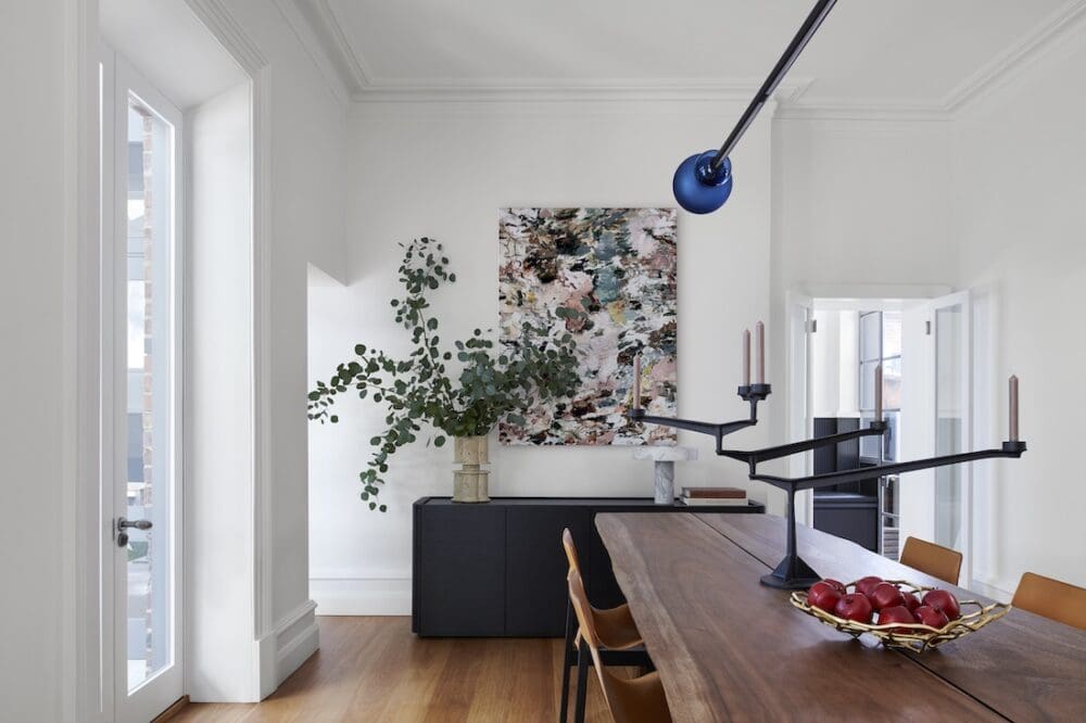 Modern dining room with timber table, sculptural black candelabra, contemporary artwork and large window bringing natural light into the minimalist space.