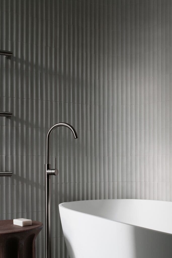 Freestanding white bathtub with brushed metal floor tap against vertically fluted grey wall tiles in a minimalist bathroom interior.  