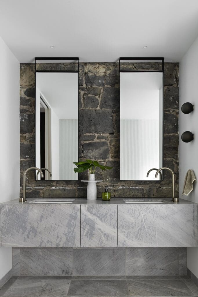 Double stone vanity with brushed metal tapware and tall mirrors against a bluestone wall in the ensuite at Bluestone Sanctuary by The Stylesmiths and YDM Studio.
