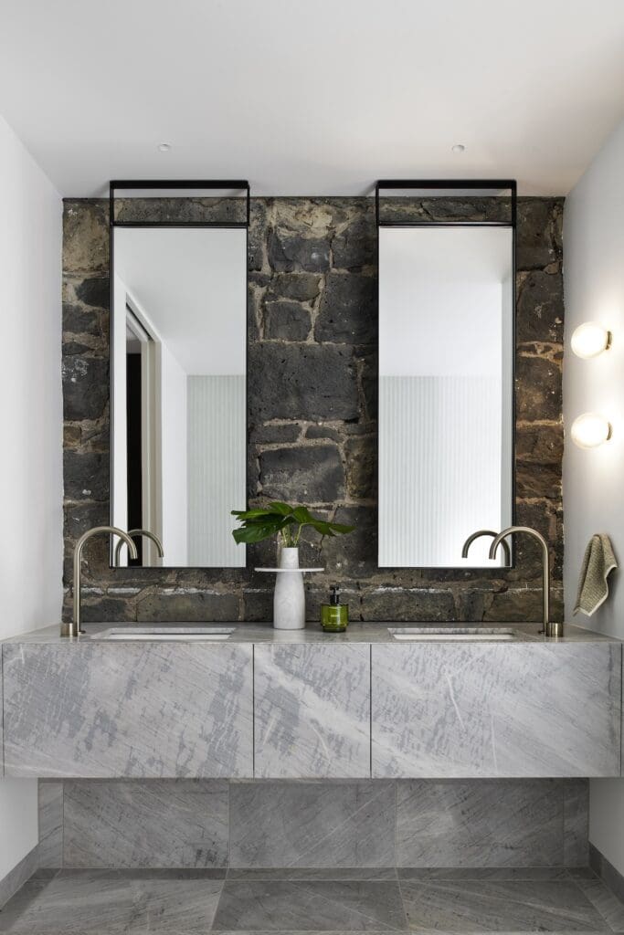 Double stone vanity with brushed metal tapware and tall mirrors against a bluestone wall in the ensuite at Bluestone Sanctuary by The Stylesmiths and YDM Studio.