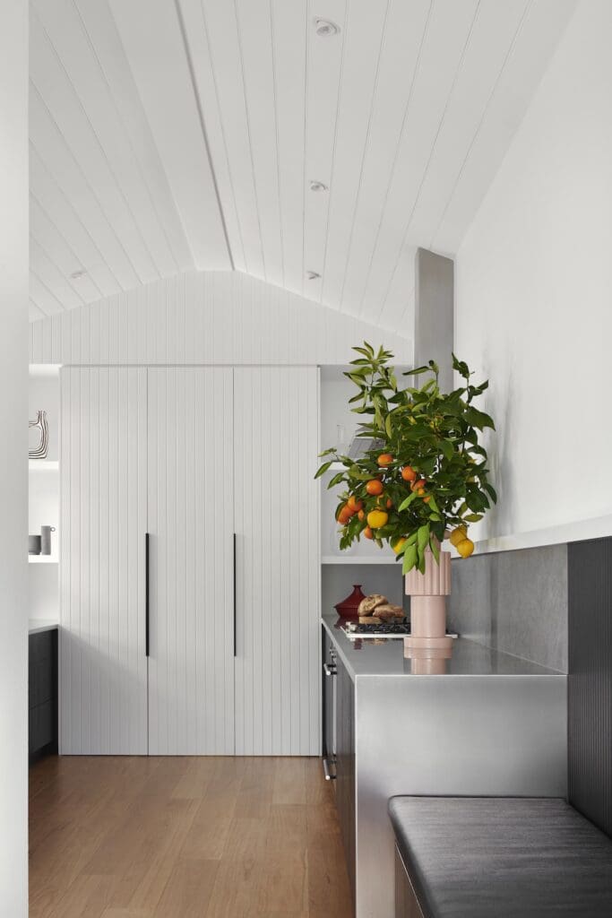 Minimalist kitchen interior at Bluestone Sanctuary featuring white V-groove cabinetry, vaulted ceiling, stainless steel bench and citrus arrangement, designed by The Stylesmiths with architecture by YDM Studio.