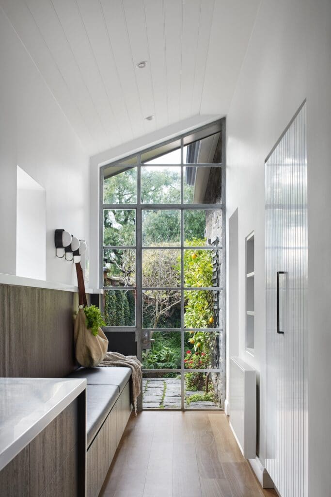 Light-filled mudroom at Bluestone Sanctuary with built-in bench seating, timber cabinetry and large grid window overlooking the garden, designed by The Stylesmiths and YDM Studio.