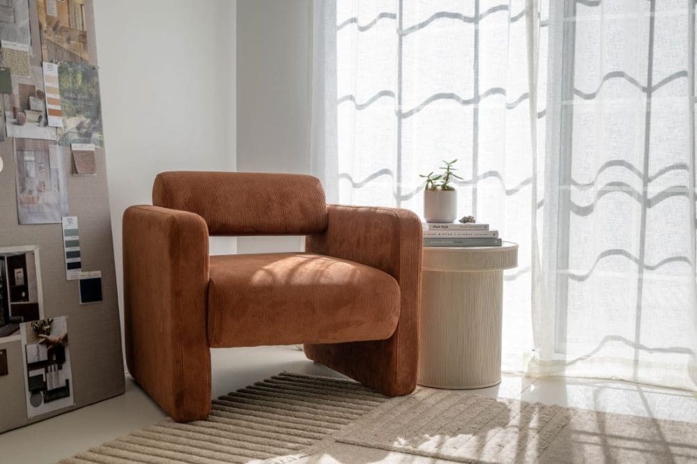 Rust upholstered armchair beside a ribbed cream side table in soft natural light.