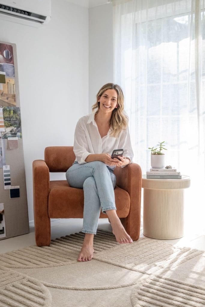 Gina Beschorner sitting in a rust armchair beside a ribbed side table in a light-filled living room.