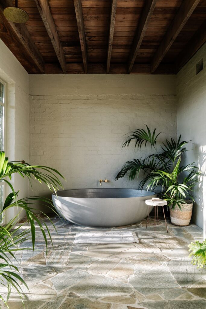 Freestanding oval bathtub beneath exposed timber ceiling beams with white brick walls, brass tapware and indoor plants in Mountain House by Breathe, photographed by Marnie Hawson.