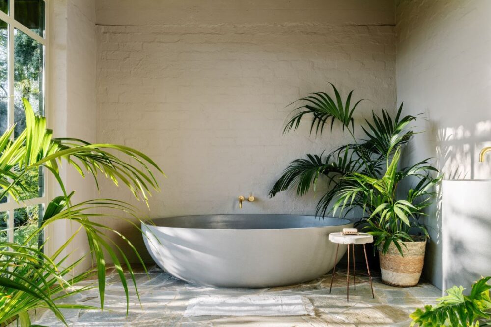 Freestanding oval bathtub set against white painted brick wall with brass tapware and lush indoor plants in Mountain House by Breathe, photographed by Marnie Hawson.
