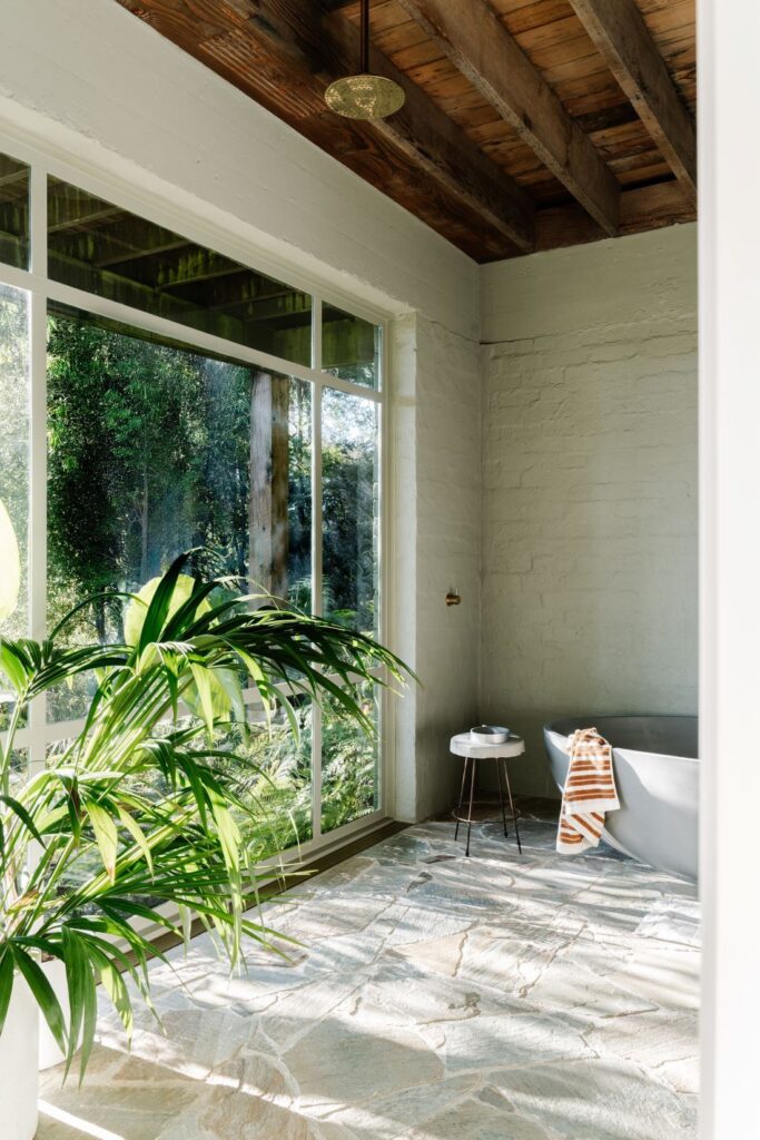 Freestanding bath beside full-height window with timber ceiling beams and natural stone flooring at Mountain House by Breathe, photographed by Marnie Hawson.