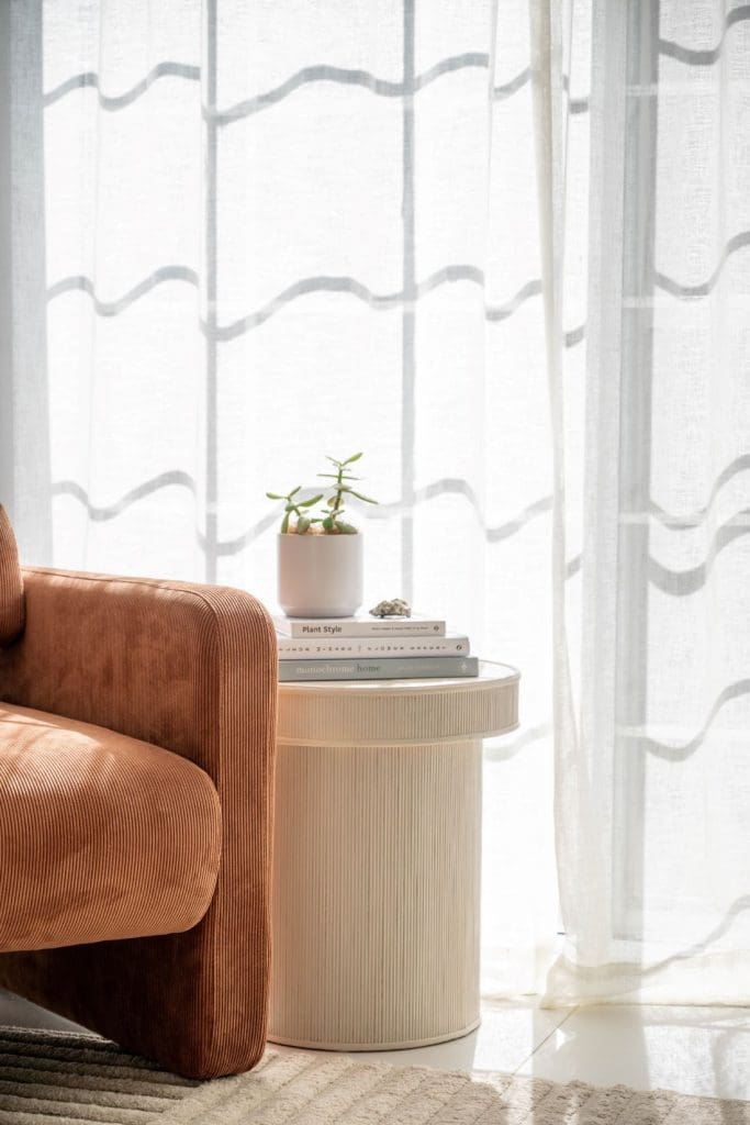 Rust velvet armchair beside a ribbed cream side table with stacked books and a small potted plant in front of sheer curtains.