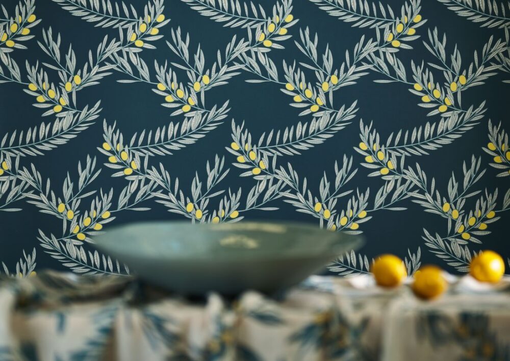 Amalfi-Lines Oliva wallpaper features a rich navy background adorned with olive branch motifs and bright yellow olives, complemented by a modern bowl and fresh lemons.