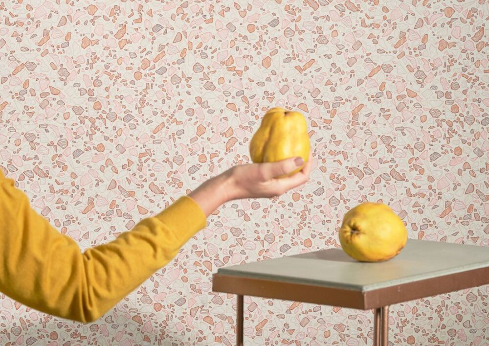 Amalfi-Lines Terrazzo wallpaper featuring soft pink and beige stone patterns, with a hand holding a quince fruit and a modern table setting.