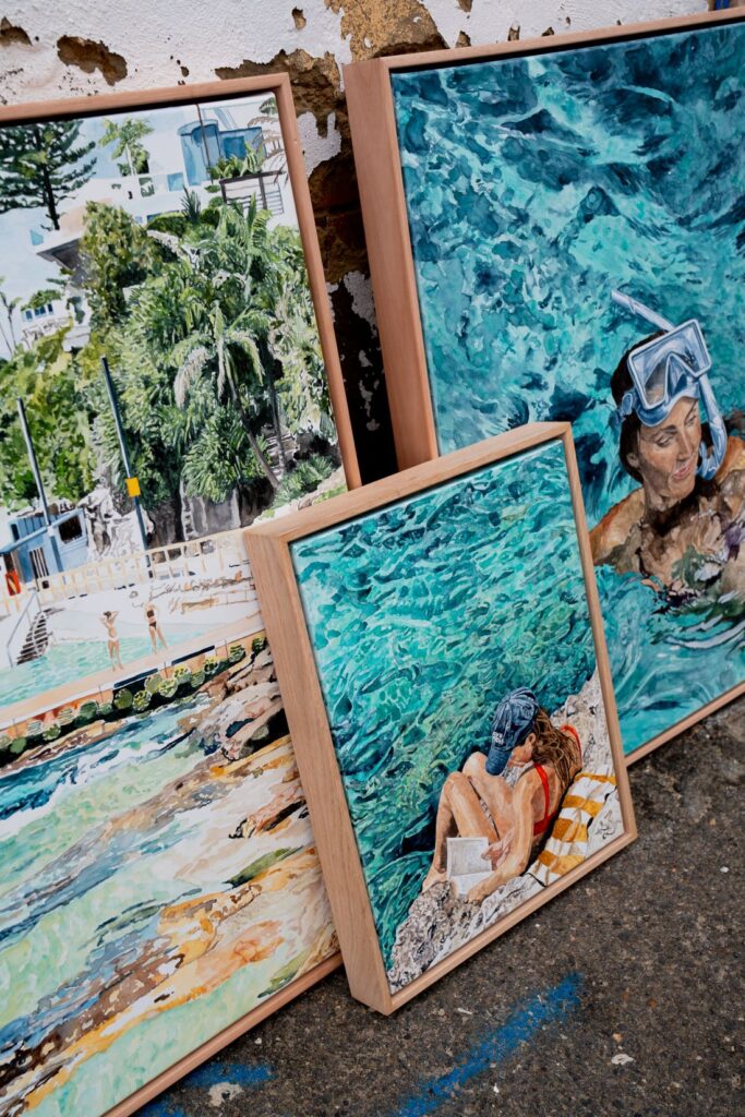 Leah Ramage’s poolside-themed artworks, featuring vibrant depictions of swimmers, lounging beachgoers, and tranquil waters, leaning against a weathered wall. The art captures moments of relaxation by the pool, showcasing the refreshing hues of clear blue water.