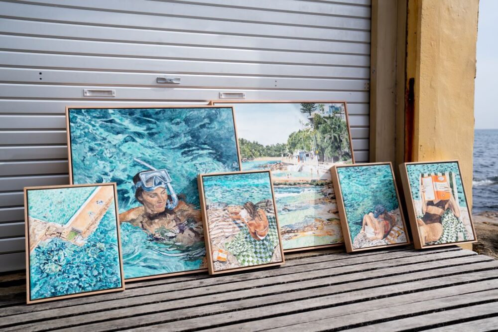 Leah Ramage’s stunning collection of poolside art displayed outdoors against a neutral backdrop. The pieces capture moments of summer relaxation, including a snorkeler, a reader by the pool, and sunbathers on striped towels, creating a serene and nostalgic atmosphere.