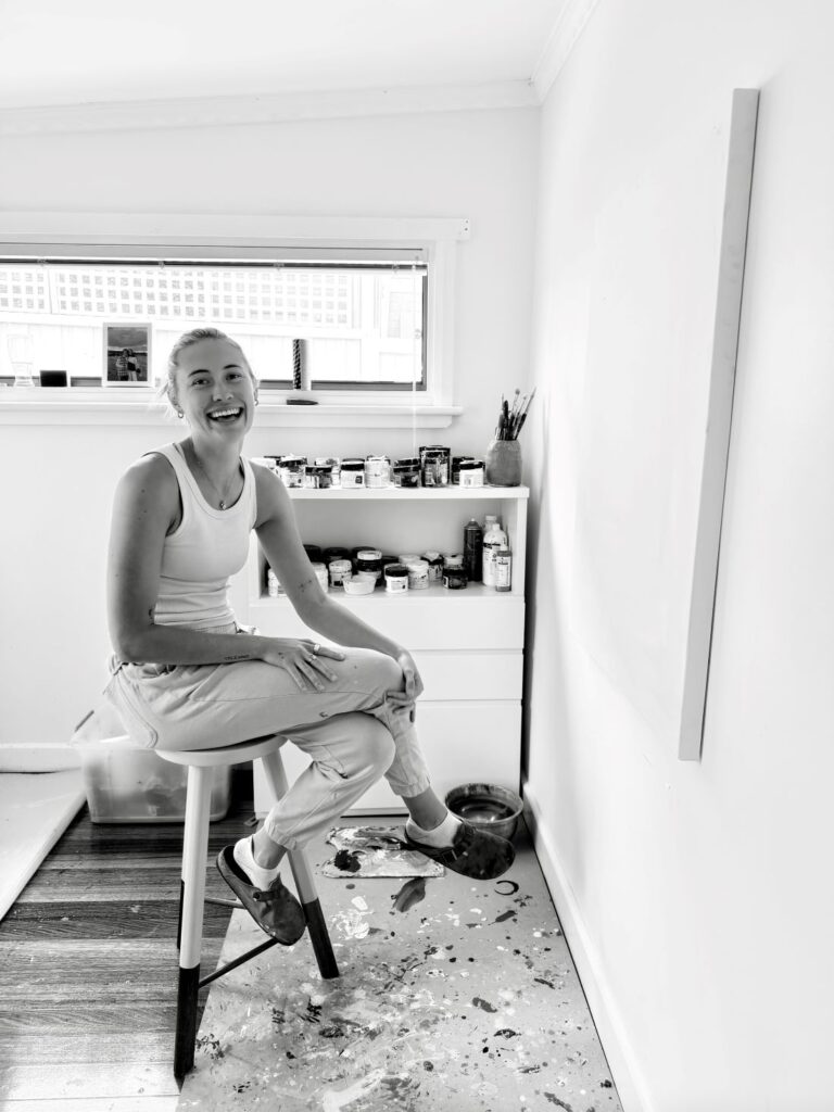 Black and white portrait of Adele Auchterlonie, a Tasmanian abstract expressionist artist, sitting on a stool in her studio surrounded by art supplies