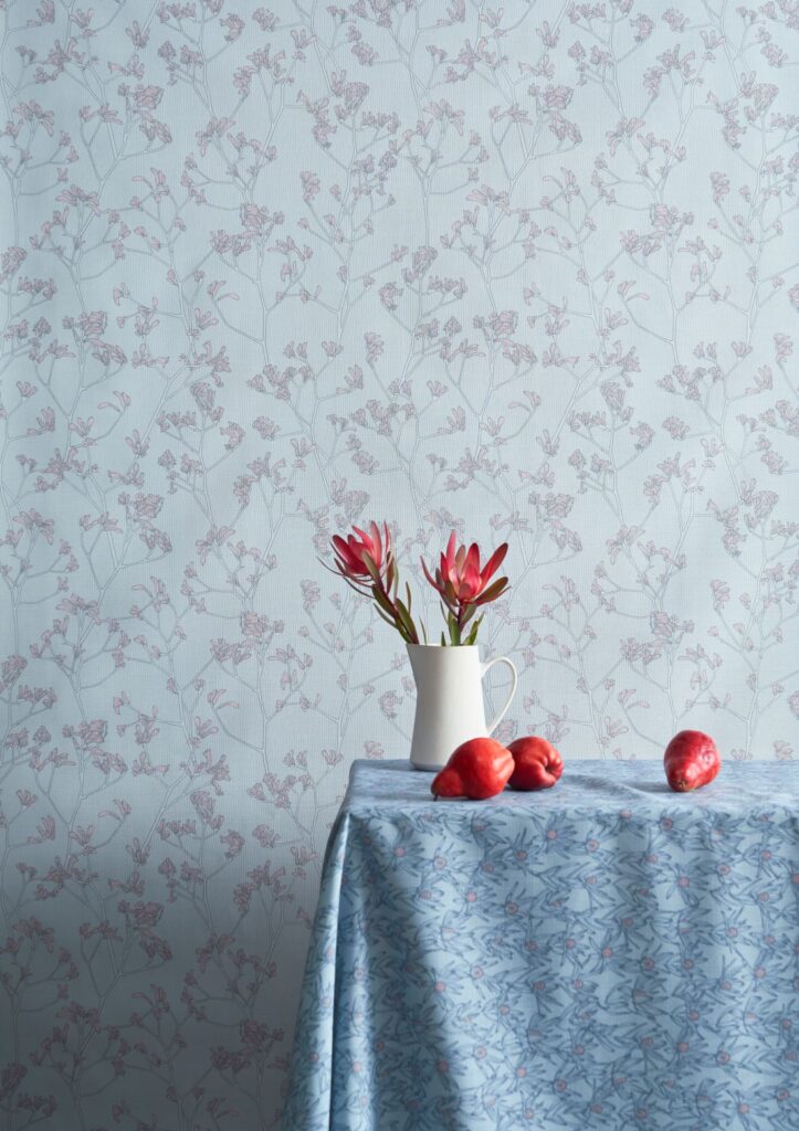 Kangaroo_Paw_ soft floral wallpaper in pastel pink and blue tones, with a table draped in a blue floral cloth, adorned with fresh flowers in a white vase and red fruit.