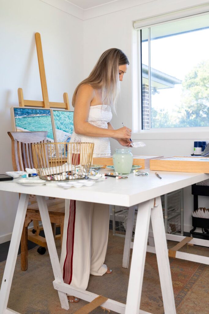 Mixing paint in their bright studio while standing at a white worktable with brushes, paint tubes, and a finished coastal artwork in the background.