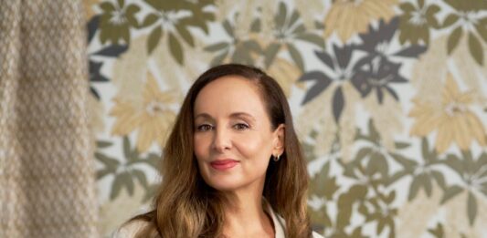 Patricia Braune, a skilled interior designer and surface artist, stands confidently in a white blazer against a botanical print wallpaper, showcasing her design expertise.