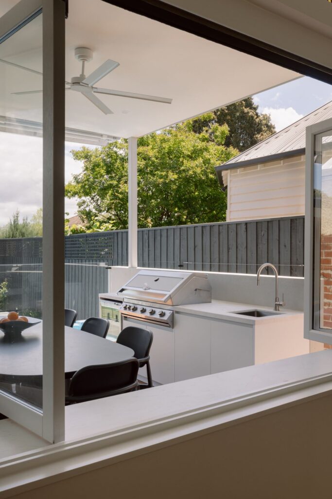 A modern outdoor BBQ area with a built-in grill, sink, and countertop, offering a clear view through large windows into the dining space