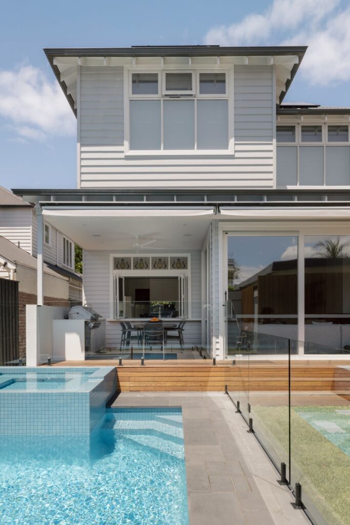 A modern outdoor area with a pool, wooden decking, and glass railings, leading to a dining space with a table and chairs