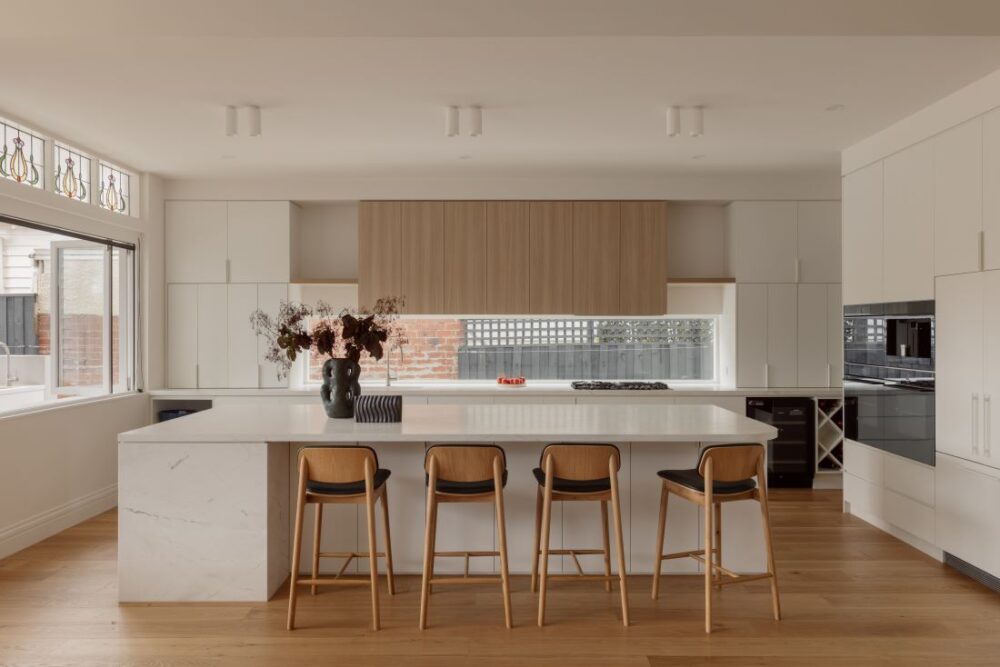 A modern kitchen with a large marble island, wooden upper cabinetry, and stylish bar stools