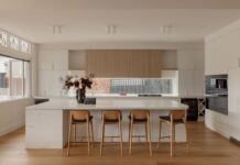 A modern kitchen with a large marble island, wooden upper cabinetry, and stylish bar stools. The space features sleek white and wood finishes.