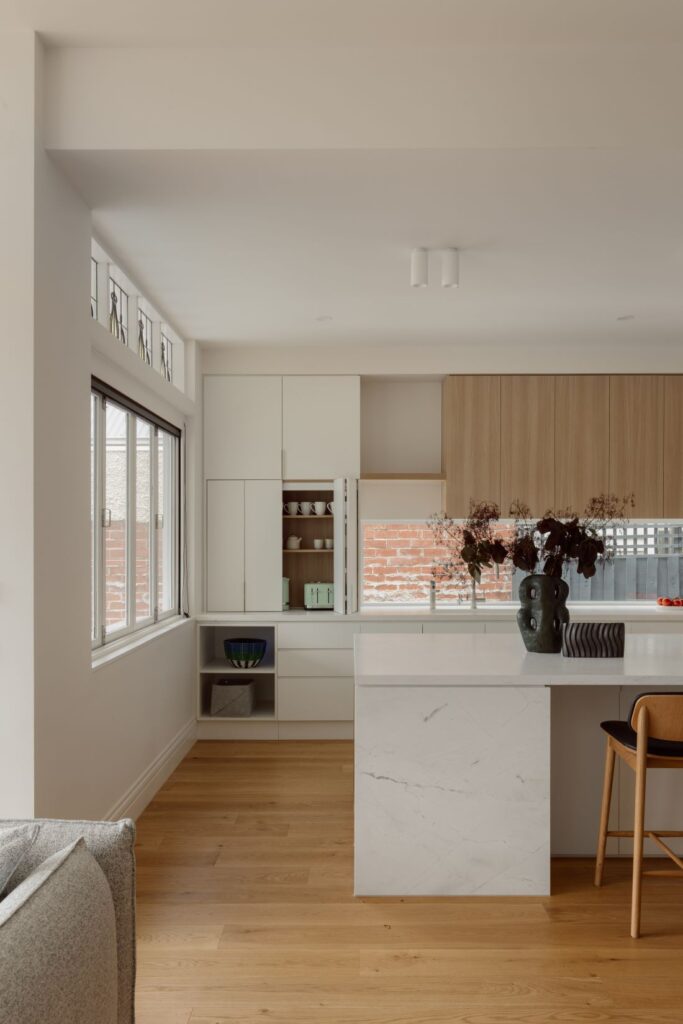 A spacious kitchen featuring a large marble island, light wooden cabinetry, and open shelving for decorative items and plants