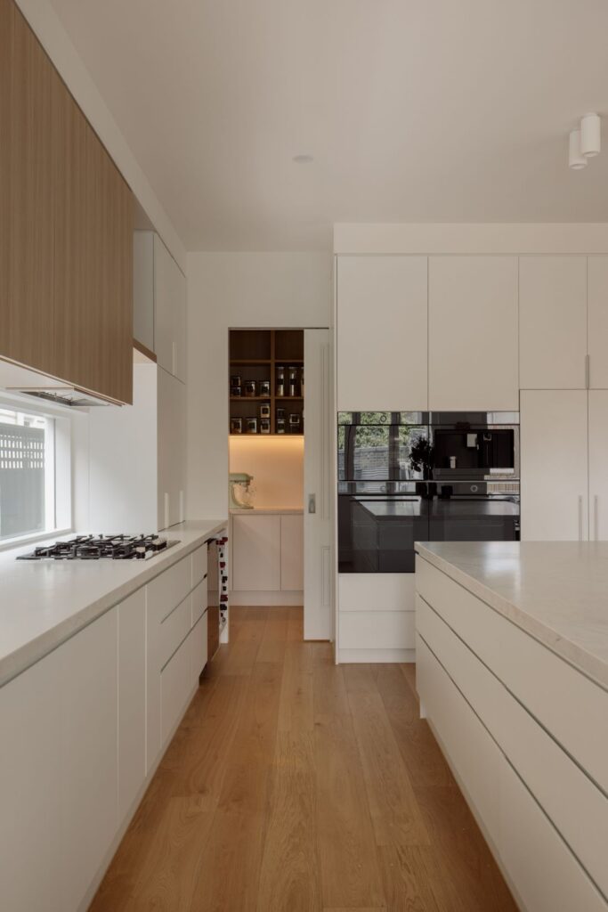 A contemporary kitchen with sleek white cabinetry, wooden accents, and hidden storage. The open doorway reveals walk in butlers pantry