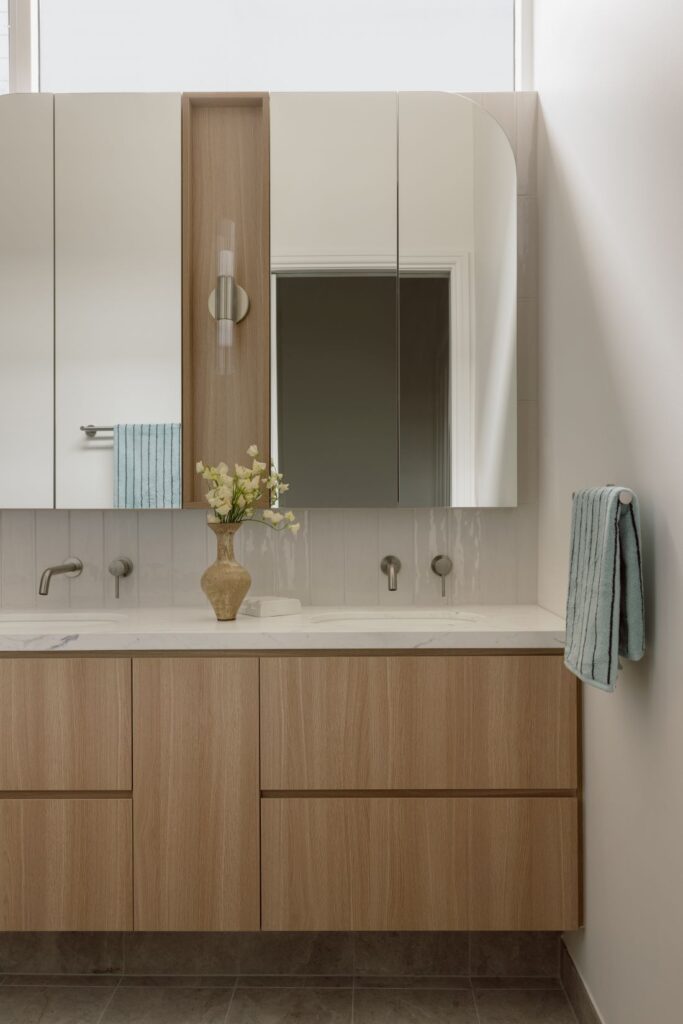 A minimalist bathroom featuring a wooden vanity, modern mirrors, and a vase with flowers, complemented by soft towels and sleek fixtures