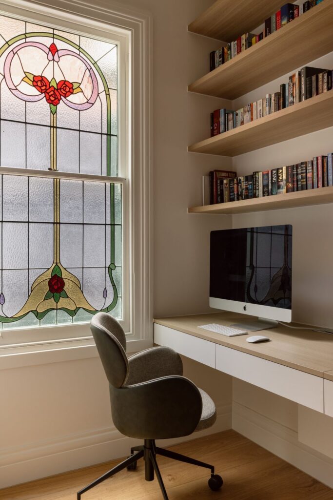 A home office featuring a modern desk with an iMac, a comfortable chair, and shelves filled with books. A stained-glass window adds color to the space.