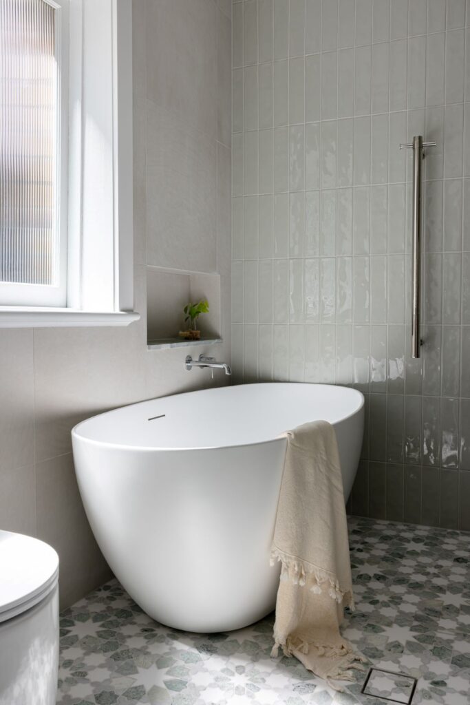 A sleek freestanding bathtub with a soft beige throw, set against a soft grey tiled wall with a recessed shelf featuring a small plant.