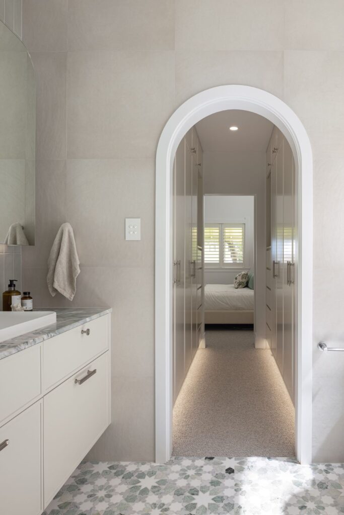 A modern bathroom with a soft beige palette, featuring a freestanding vanity, intricate patterned floor tiles, and a hallway leading to a master bedroom.