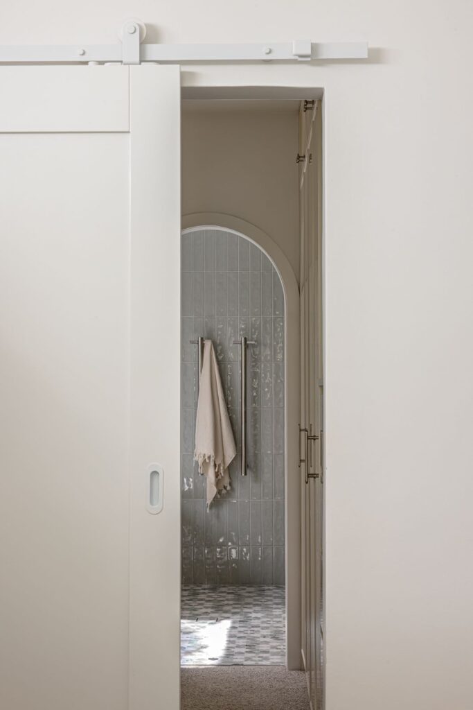 A white sliding barn door opens to a bathroom with a grey tiled shower and soft towel hanging from dual towel bars.