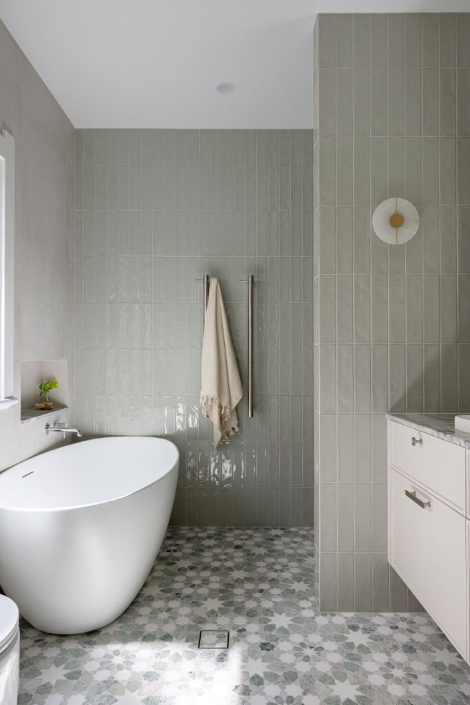 A freestanding bathtub in a bathroom with soft green tiles, a beige towel hanging on towel bars, and a patterned floor.