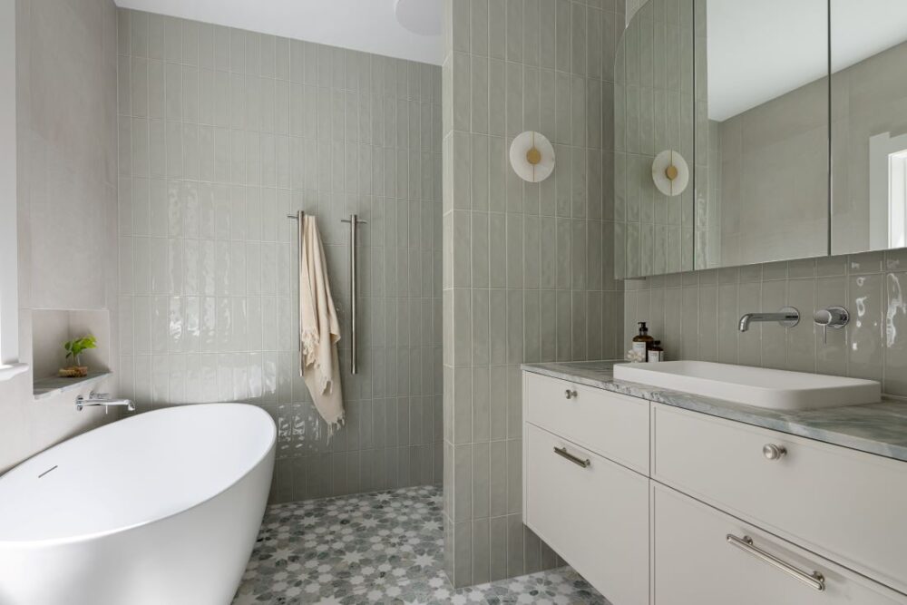 A stylish bathroom featuring a freestanding bathtub, soft green tiles, modern double sconces, and a marble-topped vanity.