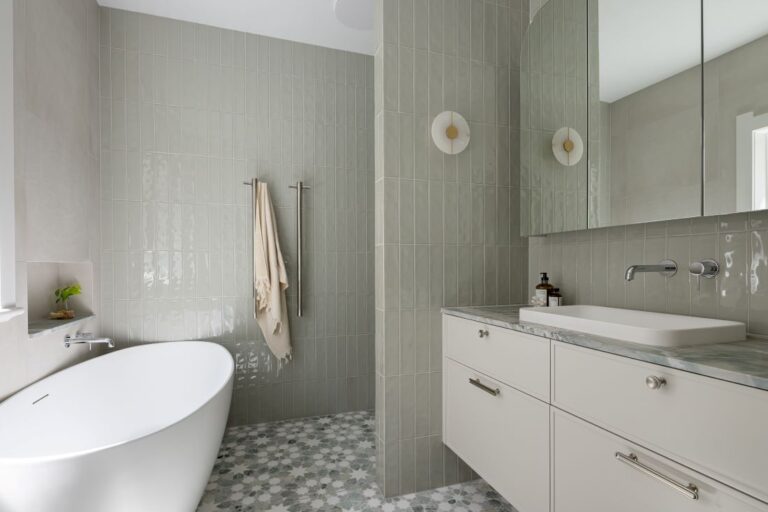 A stylish bathroom featuring a freestanding bathtub, soft green tiles, modern double sconces, and a marble-topped vanity.