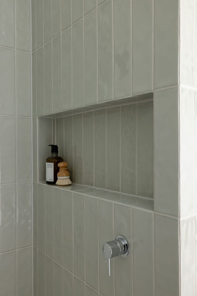A shower niche with a small bottle and natural brush on soft green tiled walls, featuring a modern shower valve.