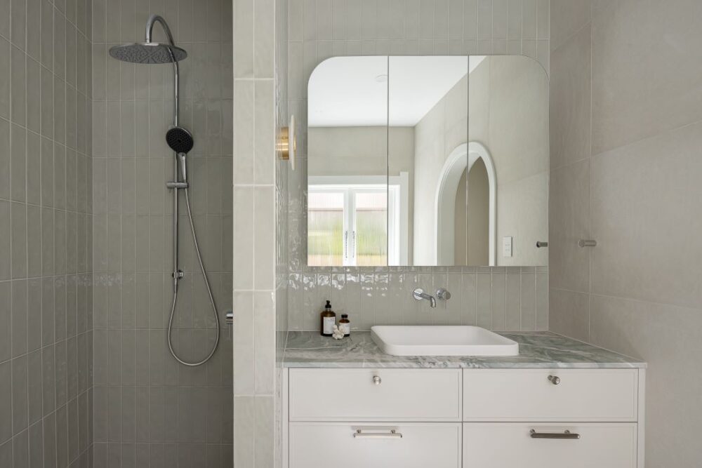 A sleek bathroom with a marble-topped vanity, overhead shower, and minimalist mirror with an arched doorway in the background.