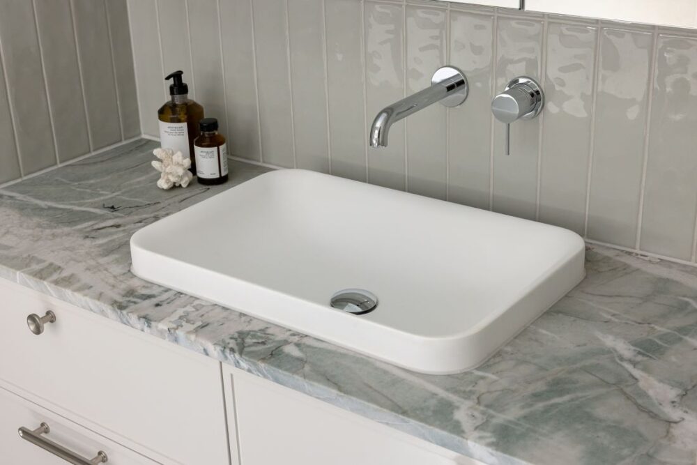 A modern white basin on a marble countertop with minimalist faucet and bath products beside it.