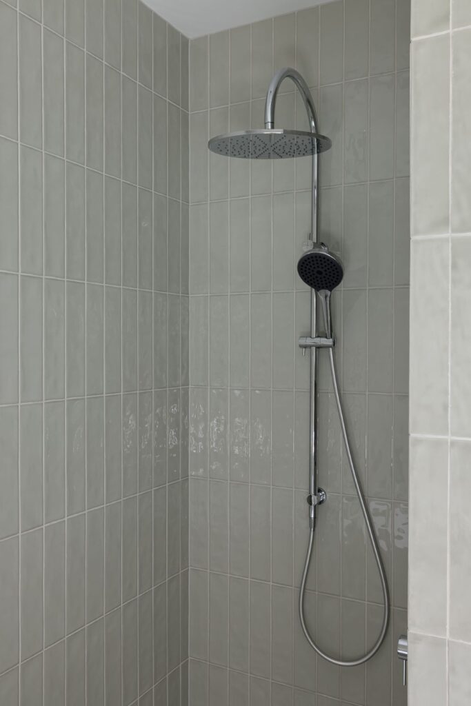 A sleek shower featuring a chrome overhead rain showerhead and handheld sprayer, set against soft green tiles.