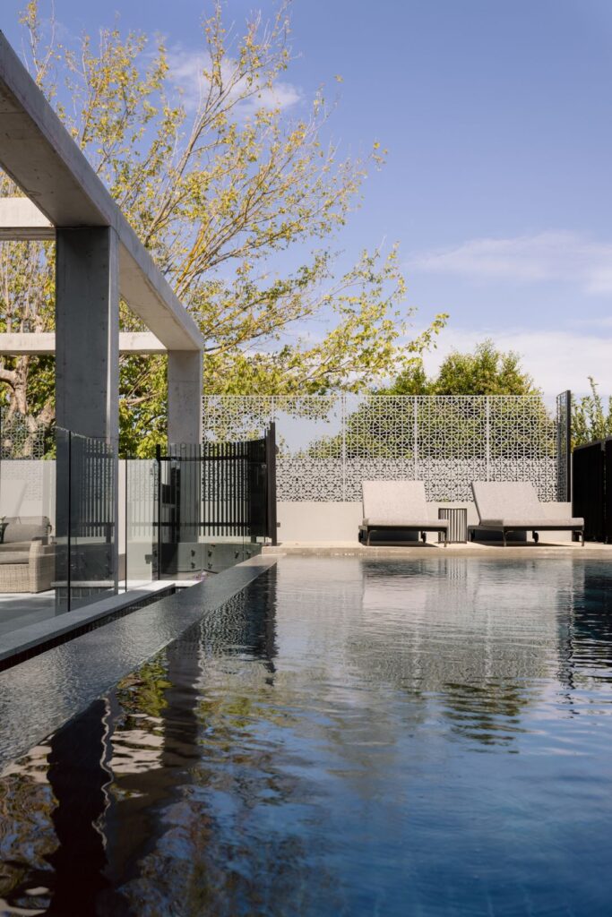 Poolside area with sleek loungers, concrete beams overhead, and greenery in the background.