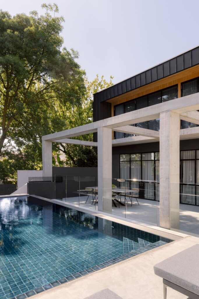 Sleek poolside with a blue-tiled pool, concrete beams overhead, and an expansive outdoor seating area.