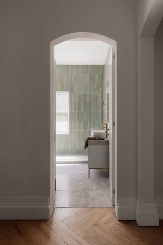 View into a bathroom with soft green tiles, minimalist vanity, and natural light.