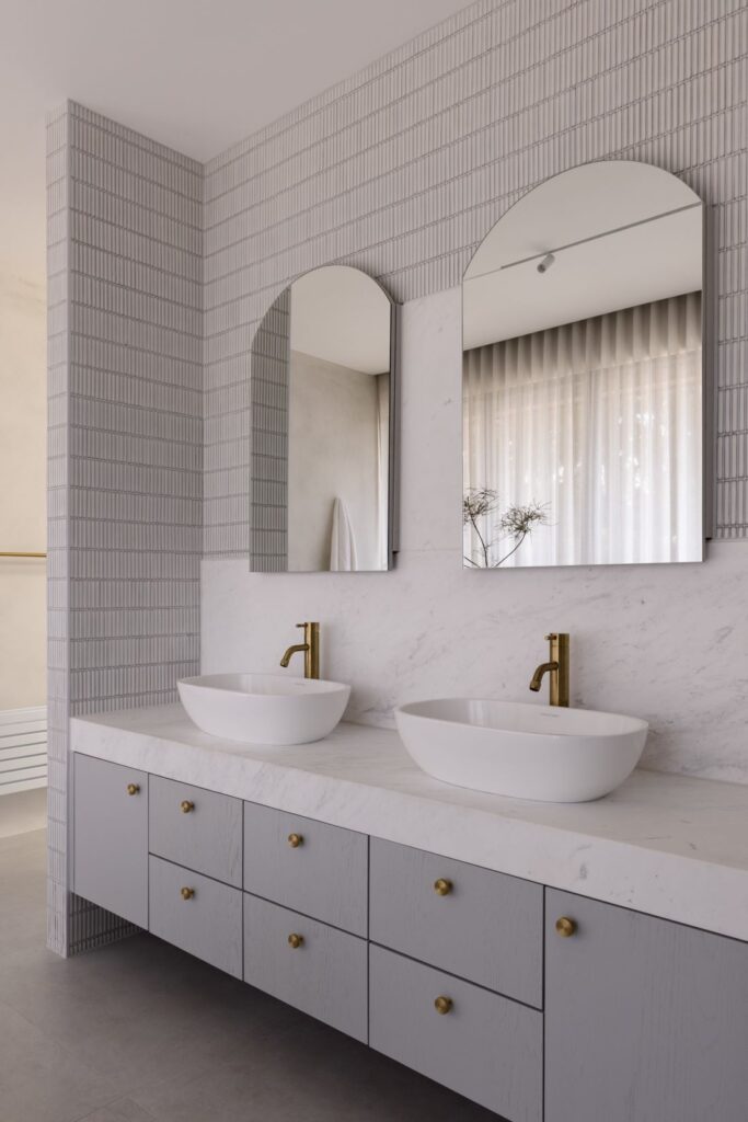 Bathroom with dual marble sinks, brass faucets, and soft grey cabinetry.