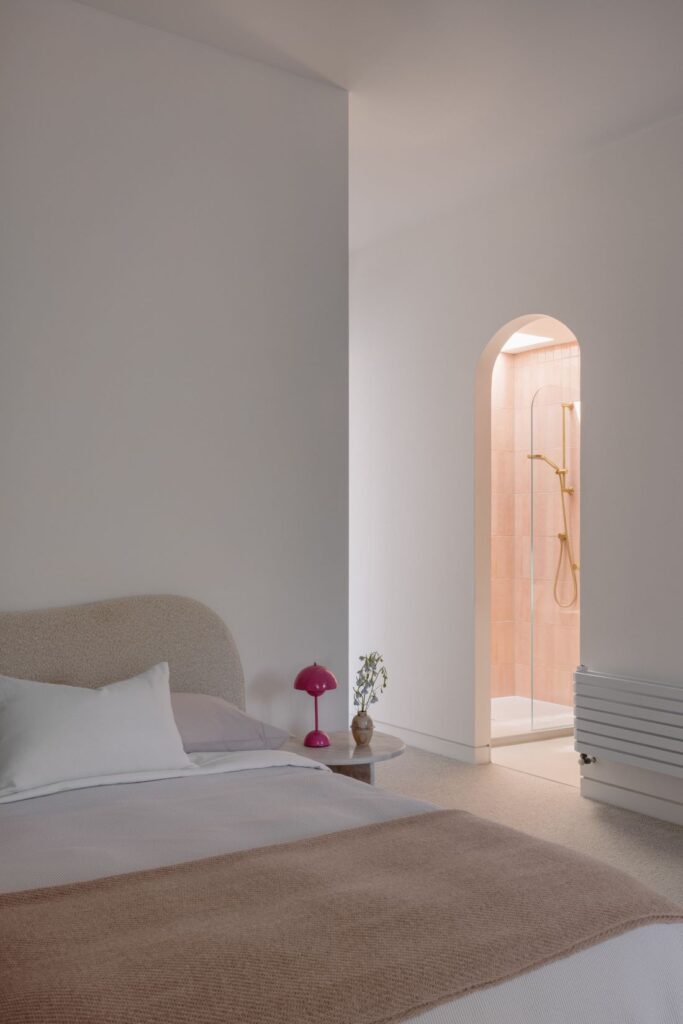 Bedroom with a minimalist bed, a playful pink lamp, and an arched doorway leading to a shower.