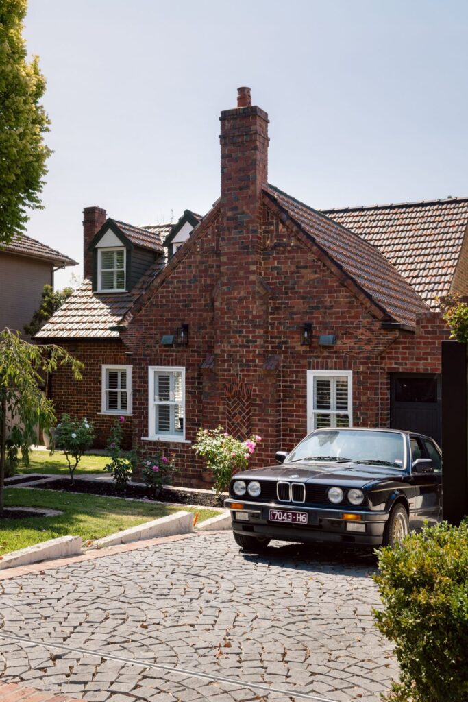Tudor-style brick home with a cobblestone driveway and a vintage BMW parked outside.