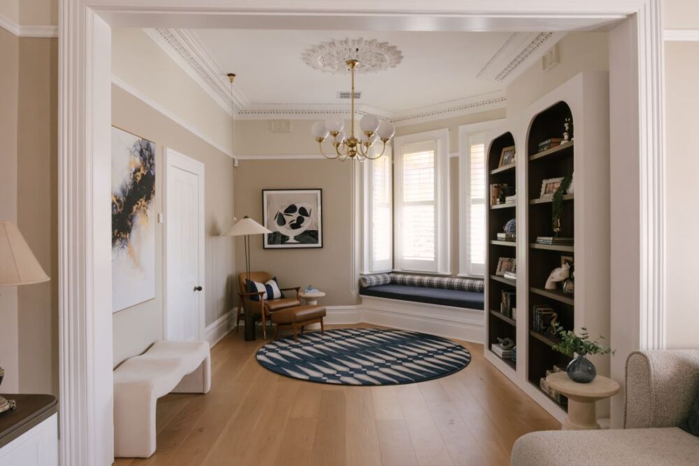 A classic formal sitting room zone at Drummoyne House - Issy & H Creative. Features a built-in bay window bench seat, a tan leather armchair with footstool, a round patterned geometric rug, custom arched bookcases, and a brass globe chandelier hanging from an ornate ceiling rose.