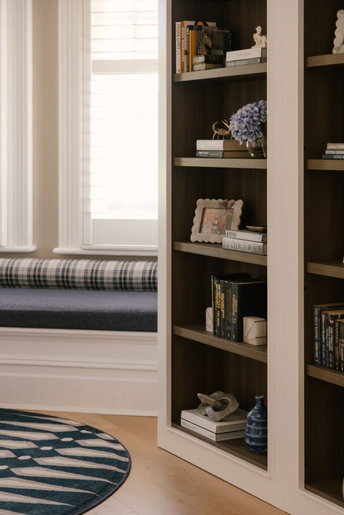 A close-up detail of a custom built-in bookcase at Drummoyne House - Issy & H Creative. The dark timber shelves are styled with curated hardcover books, a vase of blue hydrangeas, small sculptures, and a decorative picture frame, with a plaid window seat visible in the background.
