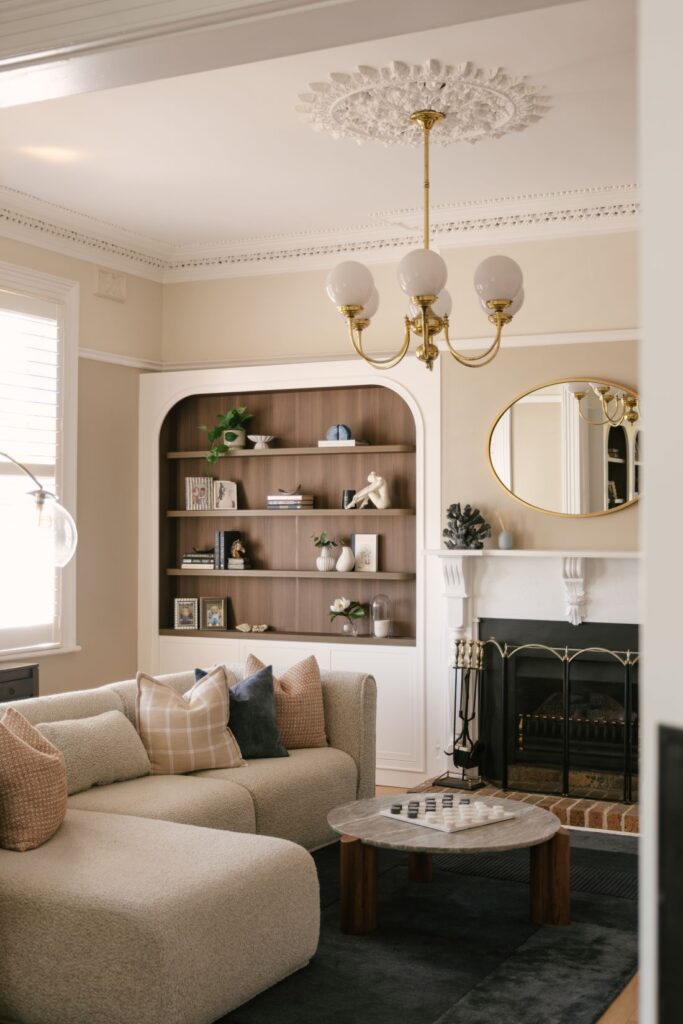 A beautifully preserved formal living room at Drummoyne House - Issy & H Creative. Showcases a modern curved bouclé sectional sofa with beige and navy pillows, a stone coffee table featuring a chess set, a built-in arched bookshelf with dark wood backing, and a classic white fireplace mantle beneath an ornate ceiling rose and brass chandelier.