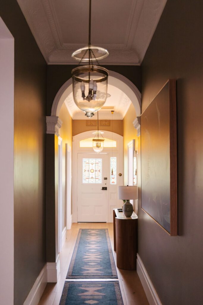 A classic long entry hallway at Drummoyne House - Issy & H Creative. Features deep olive green walls, a traditional white architectural archway, glass bell jar pendant lantern lights, a long patterned hallway runner rug, and a curved timber console table leading to a stained glass front door.