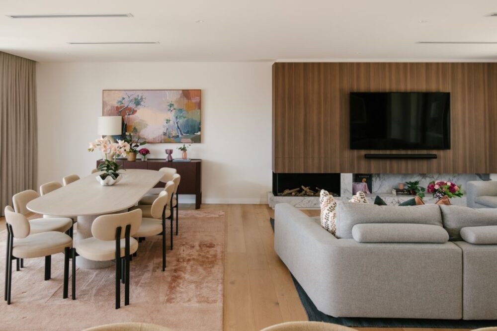 A wide interior view of the open plan dining and living space at Drummoyne House - Issy & H Creative. Showcases an oval timber dining table with cream minimalist chairs, a textured blush rug, a dark timber sideboard with abstract artwork, and a wood-panelled fireplace wall.